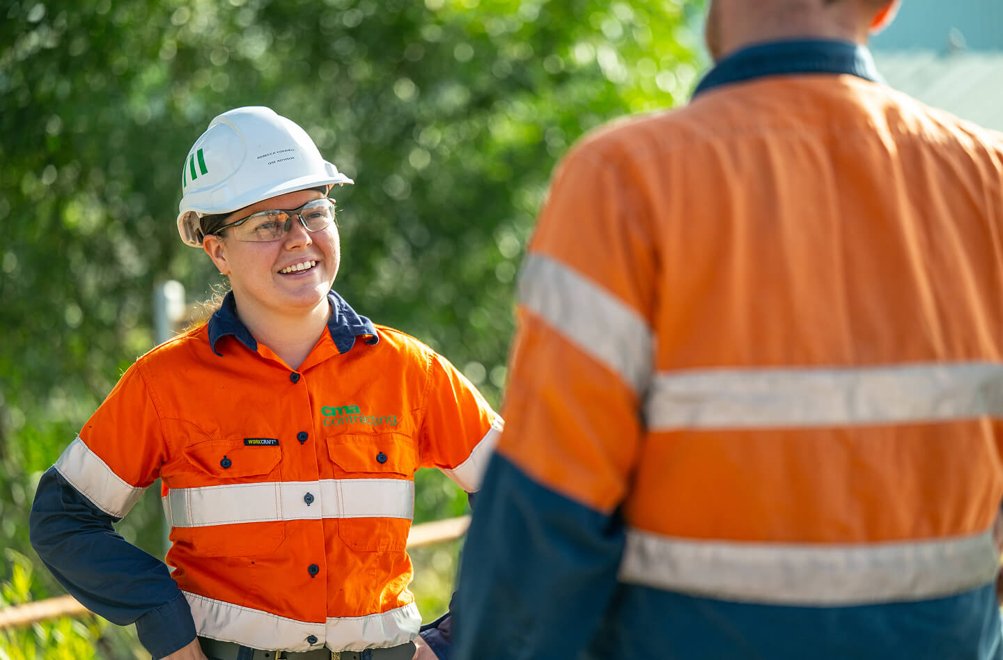 A woman wearing a hardhat and protective eyewear, talking to a person facing away from camera